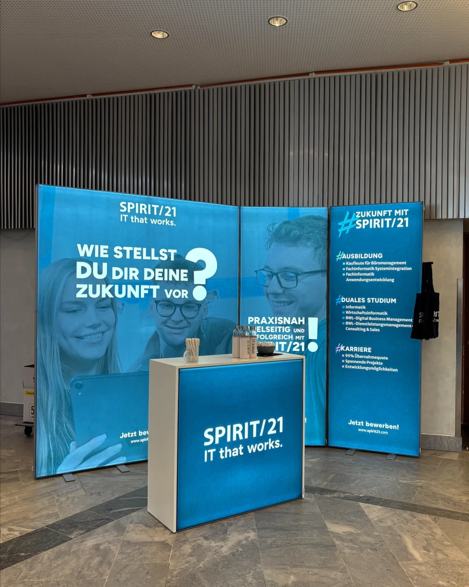 Zu sehen ist ein moderner Messestand mit vier Elementen: dem gr&ouml;&szlig;ten Element links mit den Schriftzug "Wie stellst du dir eine Zukunft vor?", links daneben zwei kleinere Elemente mit den Ausbildungs- und Studienangeboten bei SPIRIT/21. Im Vordergrund steht noch ein SPIRIT/21-gebrandeter Tresen | SPIRIT/21