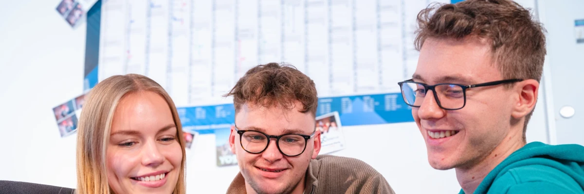 Three young people are looking at an iPad: a blonde white woman on the left and two dark blonde men with glasses in the middle and on the right. In the background you can see a large wall calendar | SPIRIT/21