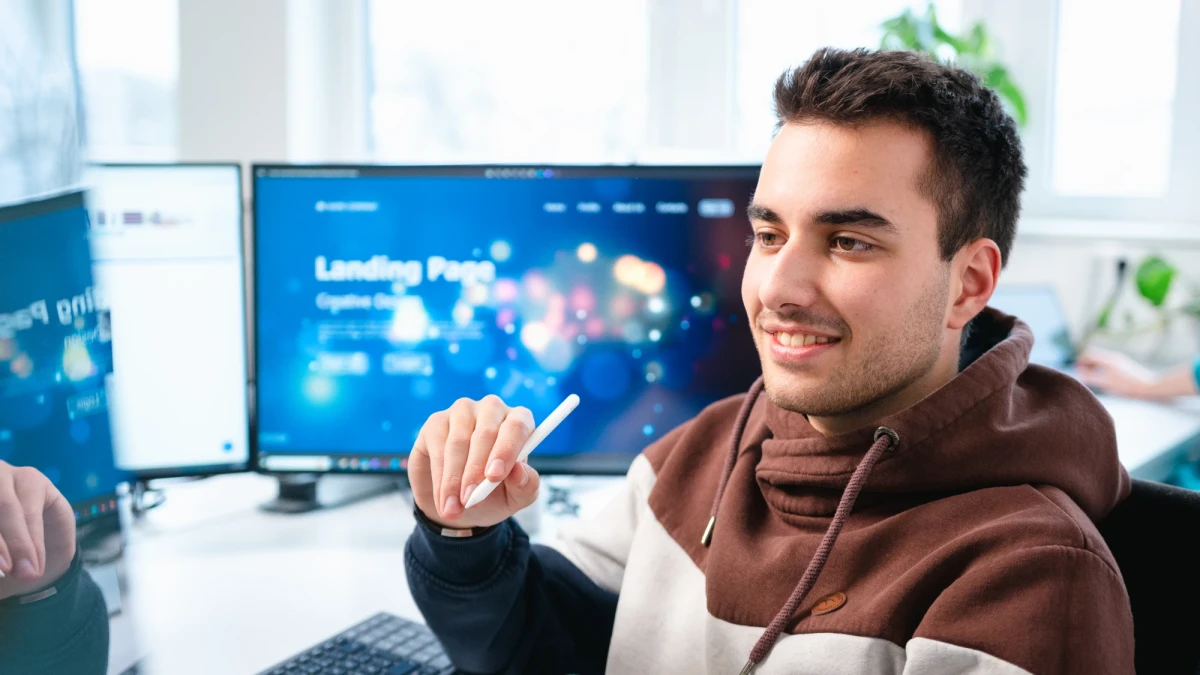Ein junger Mann mit dunklen Haaren sitzt auf einem B&uuml;rostuhl. In der Hand h&auml;lt er einen Stit f&uuml;r ein Tablet. Im Hintergrund sieht man Monnitore. | SPIRIT/21