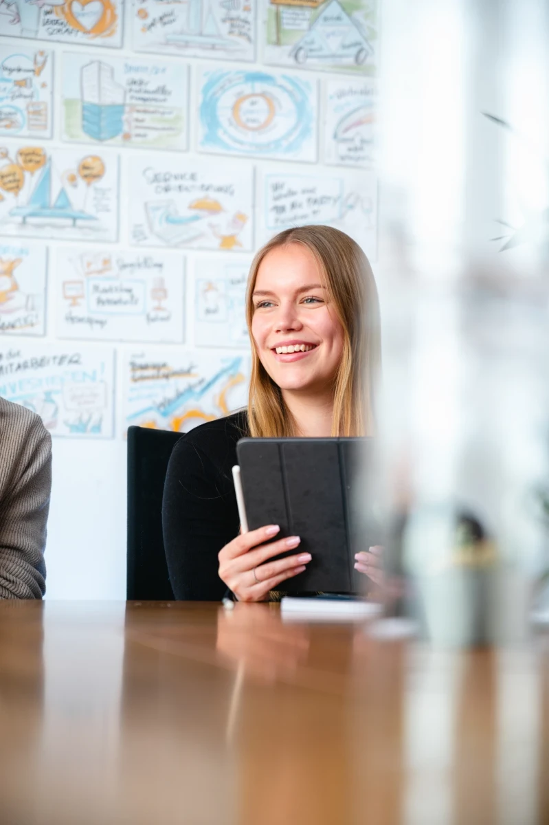 Eine junge blode Frau sitzt in einem Besprechungsraum und lacht im Gespr&auml;ch mit Ihren Kolleginnen und Kollegen | SPIRIT/21