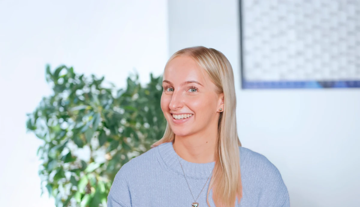 Eine junge Frau lacht in die Kamera. Sie ist in einem B&uuml;ro, im Hintergrund ist ein Wandkalender und eine Pflanze sichtbar. | SPIRIT/21