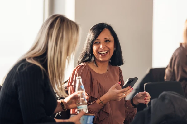 Eine Frau mit dunklen Haaren unterhält sich mit einer blonden Frau, die man nur von hinten sieht, und lächelt. Dabei hält sie ein Smartphone in der Hand | SPIRIT/21