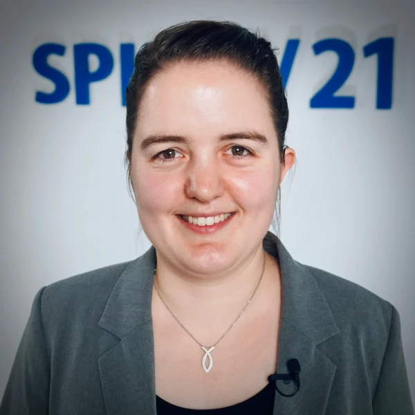 Portraitfoto einer jungen Frau mit dunklen Haaren (im Zopf) und braunen Augen, die in die Kamera l&auml;chelt. Im Hintergrund ist das SPIRIT/21-Logo | SPIRIT/21