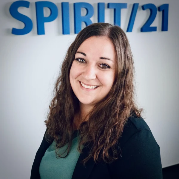 Eine junge Frau mit langen braunen welligen Haaren und braunen Augen l&auml;chelt in die Kamera. Im Hintergrund ist das SPIRIT/21-Logo zu sehen | SPIRIT/21