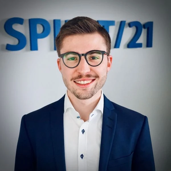 Portraitfoto eines jungen Mannes mit dunklen Haaren, dunklen Augen und Brille, der in die Kamera l&auml;chelnt. Im Hintergrund ist das SPIRIT/21-Logo | SPIRIT/21