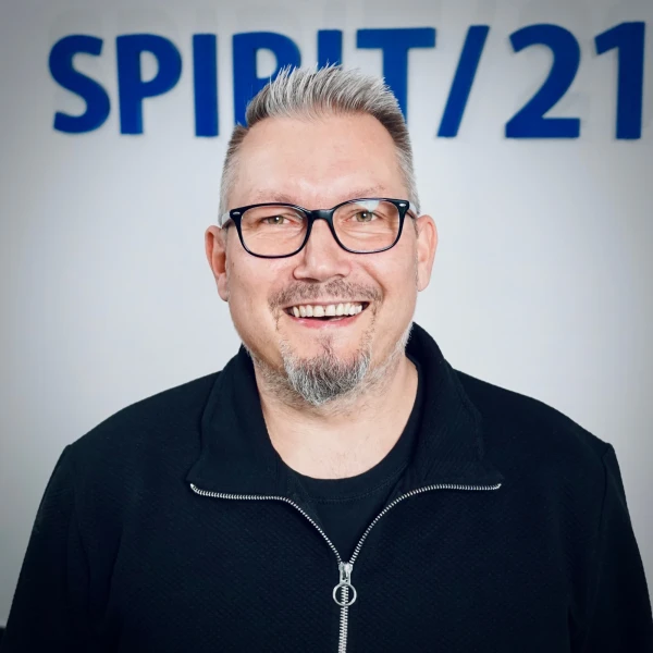 Portraitfoto eines mittelalten Mannes mit gr&auml;ulichen Haaren und einer Brille, der herzlich in die Kamera lacht. Im Hintergrund ist das SPIRIT/21-Logo | SPIRIT/21