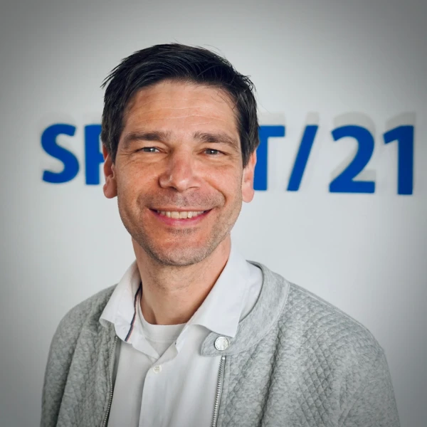 Portraitfoto eines mittelalten Mannes mit dunklen Haaren. Er tr&auml;gt ein wei&szlig;es Hemd und eine graue Jacke dar&uuml;ber und l&auml;chelt direkt in die Kamera. Im Hintergrund ist das SPIRIT/21-Logo | SPIRIT/21