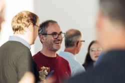 In der Mitte steht ein l&auml;chelnder mittelalter Mann mit Brille und rotem, d&uuml;nnen Pullover. Im Vorder- und Hintergrund sind weitere Personen | SPIRIT/21