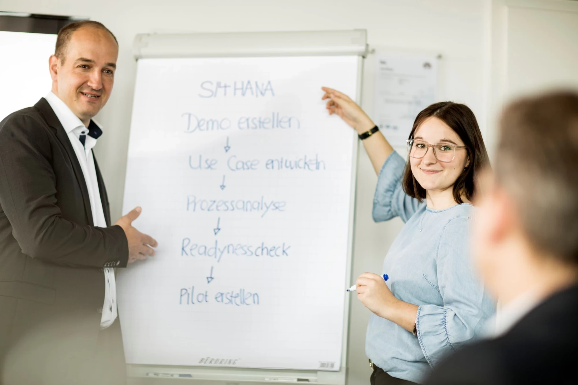 Ein Mann (links) und eine Frau (rechts) stehen vor einem Whiteboard und pr&auml;sentieren in gesch&auml;ftlichen Bereich das Thema SAP S/4HANA | SPIRIT/21