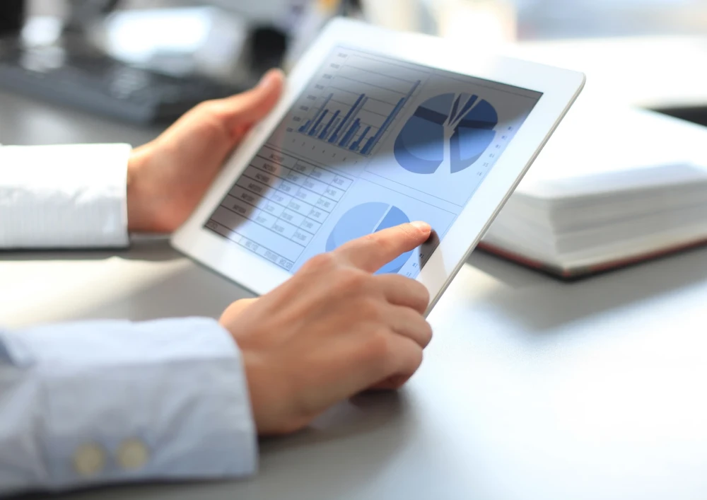 Two Hands holding an IPad with different Diagramms on it. In the background you can see different office tools.